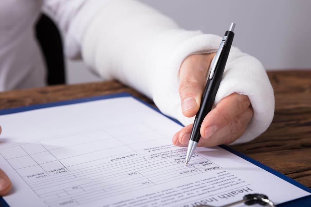 Close-up of a person with a broken arm completing a bodily injury claim form on a wooden desk.